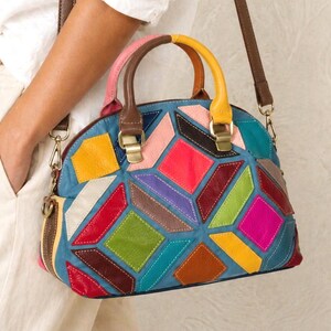 Könnte beinhalten: Eine kuppelförmige Handtasche mit einem mehrfarbigen geometrischen Patchwork-Design. Die Tasche besteht aus verschiedenen lederartigen Paneelen in Blau-, Rot-, Gelb-, Grün-, Rosa- und Brauntönen, mit zwei Tragegriffen und einem abnehmbaren Schulterriemen.