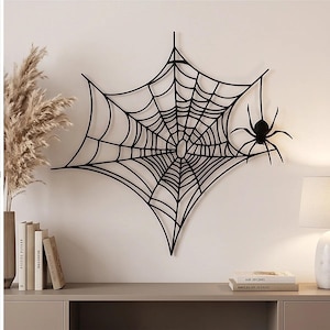 May include: A black metal wall art piece shaped like a spider web with a spider attached to the right side. The web features a central circular opening with radiating lines and concentric rings, creating a geometric pattern on the wall.