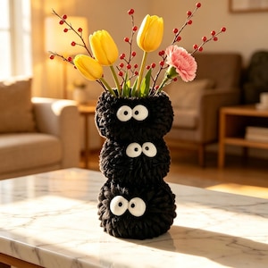 May include: A quirky black vase with three stacked sections, each with a pair of white, cartoon eyes. The vase displays yellow tulips, pink carnations, and red berries, forming a colourful floral arrangement on a marble table.