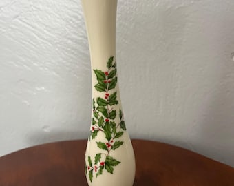 May include: A tall, slender, cream-coloured ceramic bud vase featuring a delicate holly pattern with green leaves and bright red berries. The vase has a flared base and a narrow neck, accented with a thin gold rim at the top and base.