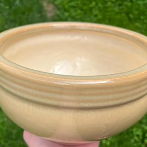 May include: A ceramic bowl with a light tan glaze. The bowl has a rounded shape with a slightly flared rim and horizontal ridges around the exterior. The interior is a lighter shade of tan. The bowl is likely used for serving food.