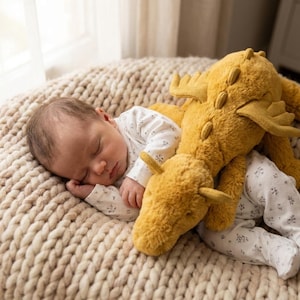 May include: A sleeping baby in a white floral onesie lies on a beige knitted surface, with a mustard-yellow dragon plush toy. The dragon has wings and a long neck. The background is softly lit, creating a gentle atmosphere.
