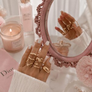 May include: A hand wearing multiple gold rings with unique designs, including a thick band, a wavy design, and a ring with small spheres. The hand is reflected in a pink framed mirror. A lit candle, perfume bottle, and a pink book with the word "Dior" are also visible.