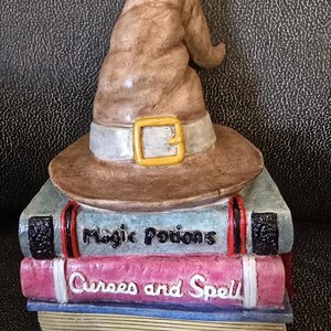 Puede incluir: Figura decorativa con un sombrero de bruja marrón con hebilla dorada, sobre una pila de libros. Los libros llevan las inscripciones "Magic Potions" y "Curses and Spell" en blanco, con detalles rojos y negros.