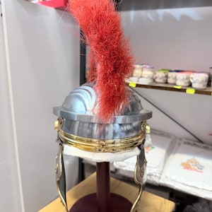 Puede incluir: Un casco de estilo romano con una c&uacute;pula de metal plateado pulido, una banda ancha de color lat&oacute;n alrededor de la base y dos protectores de mejillas articulados. Un penacho de cepillo rojo brillante, alto y prominente, se extiende verticalmente.