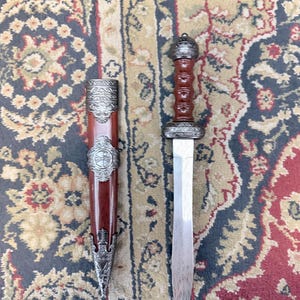 Pu&ograve; includere: Una spada gladius con lama in acciaio a doppio taglio con un sottile motivo ondulato, un'impugnatura marrone scanalata e un pomolo argentato decorato. Il fodero abbinato &egrave; marrone con intricate finiture in metallo argentato.