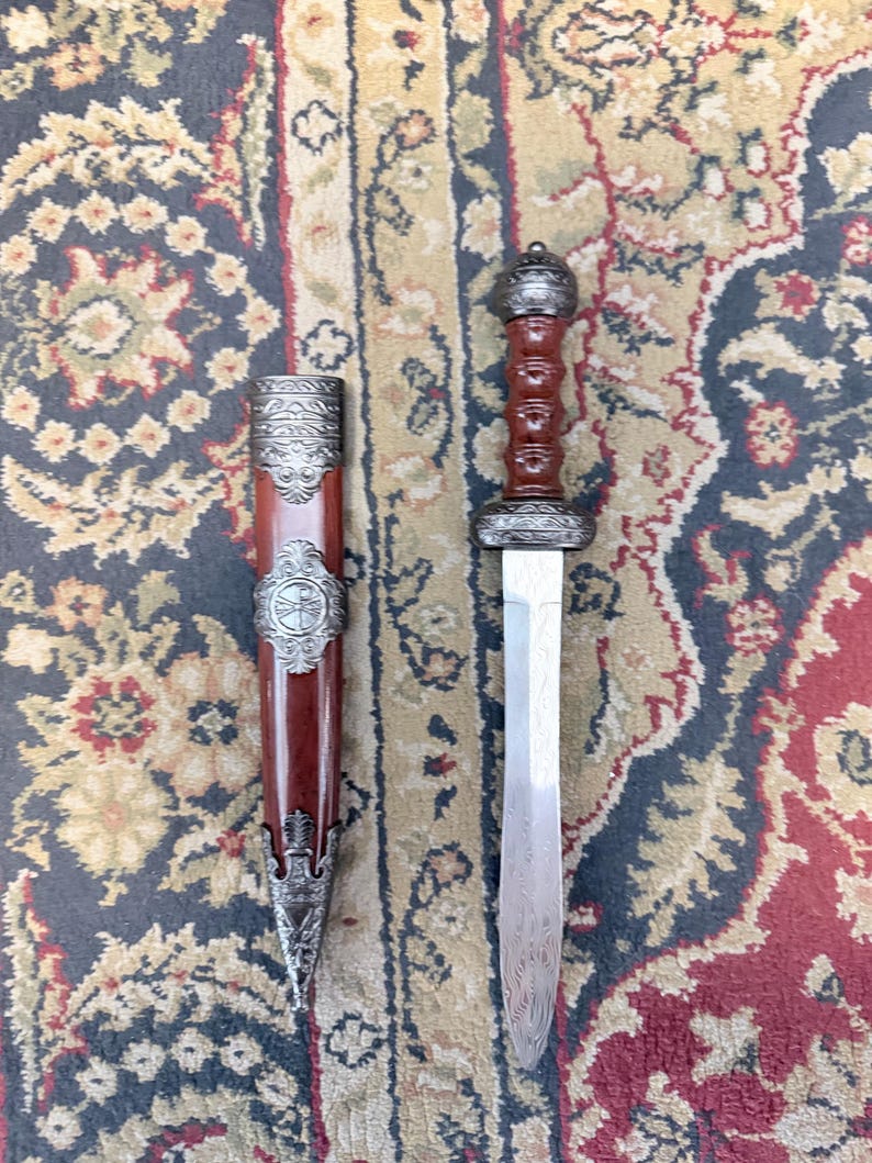 Pu&ograve; includere: Una spada gladius con lama in acciaio a doppio taglio con un sottile motivo ondulato, un'impugnatura marrone scanalata e un pomolo argentato decorato. Il fodero abbinato &egrave; marrone con intricate finiture in metallo argentato.