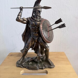 以下が含まれることがあります： 兜、マント、鎧を身に着けたスパルタの戦士のブロンズ調の彫像。右手には槍を持ち、左手には2本の矢が刺さった円形の盾を持っている。台座には「LEONIDAS」という名前が浮き彫りにされている。