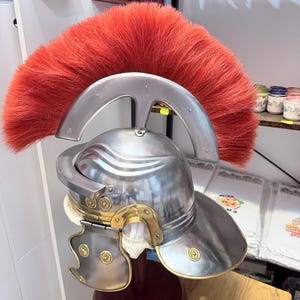 Puede incluir: Un casco de estilo romano de metal plateado con una cresta ancha y prominente de color rojo brillante hecha de pelo grueso. El casco incluye detalles en color lat&oacute;n, tachuelas decorativas circulares y carrilleras articuladas sobre un soporte.