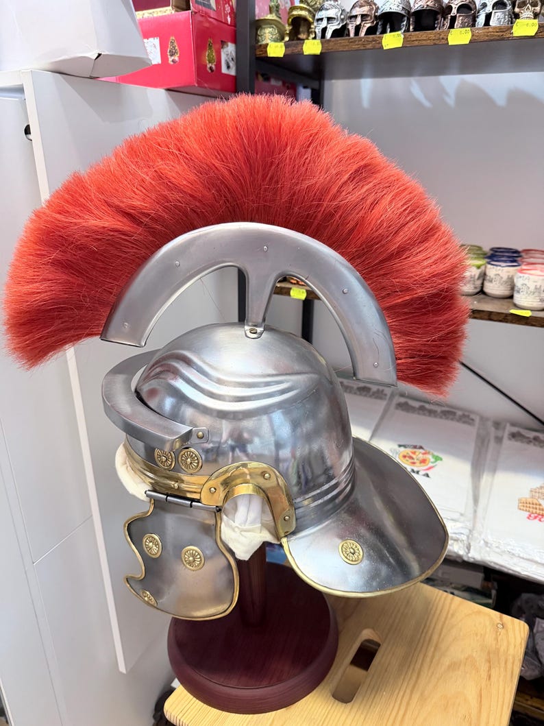 Puede incluir: Un casco de estilo romano de metal plateado con una cresta ancha y prominente de color rojo brillante hecha de pelo grueso. El casco incluye detalles en color lat&oacute;n, tachuelas decorativas circulares y carrilleras articuladas sobre un soporte.