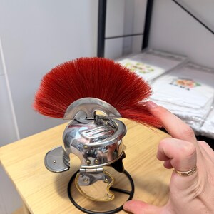 K&ouml;nnte beinhalten: Ein Miniaturhelm aus silberfarbenem Metall mit einem markanten, f&auml;cherf&ouml;rmigen Kamm aus steifen, leuchtend roten Borsten. Der Helm verf&uuml;gt &uuml;ber eine abgerundete Kuppel, einen geschwungenen Nackenschutz und bewegliche Wangenklappen mit goldenen Akzenten.
