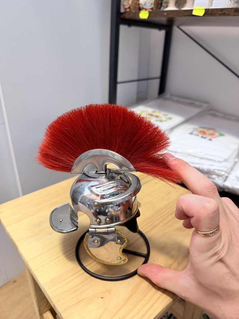 K&ouml;nnte beinhalten: Ein Miniaturhelm aus silberfarbenem Metall mit einem markanten, f&auml;cherf&ouml;rmigen Kamm aus steifen, leuchtend roten Borsten. Der Helm verf&uuml;gt &uuml;ber eine abgerundete Kuppel, einen geschwungenen Nackenschutz und bewegliche Wangenklappen mit goldenen Akzenten.