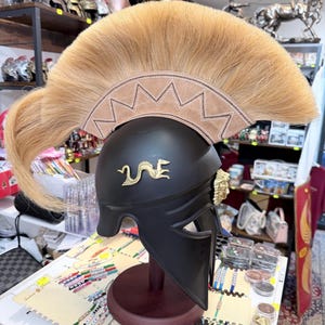 K&ouml;nnte beinhalten: Ein mattschwarzer Helm mit einem markanten, beigefarbenen Rosshaarkamm auf einer Basis mit Zickzack-N&auml;hten. Der Helm ist an der Seite mit einem goldenen Drachenemblem und in der N&auml;he des Gesichtsschutzes mit einem goldenen L&ouml;wenkopf verziert.