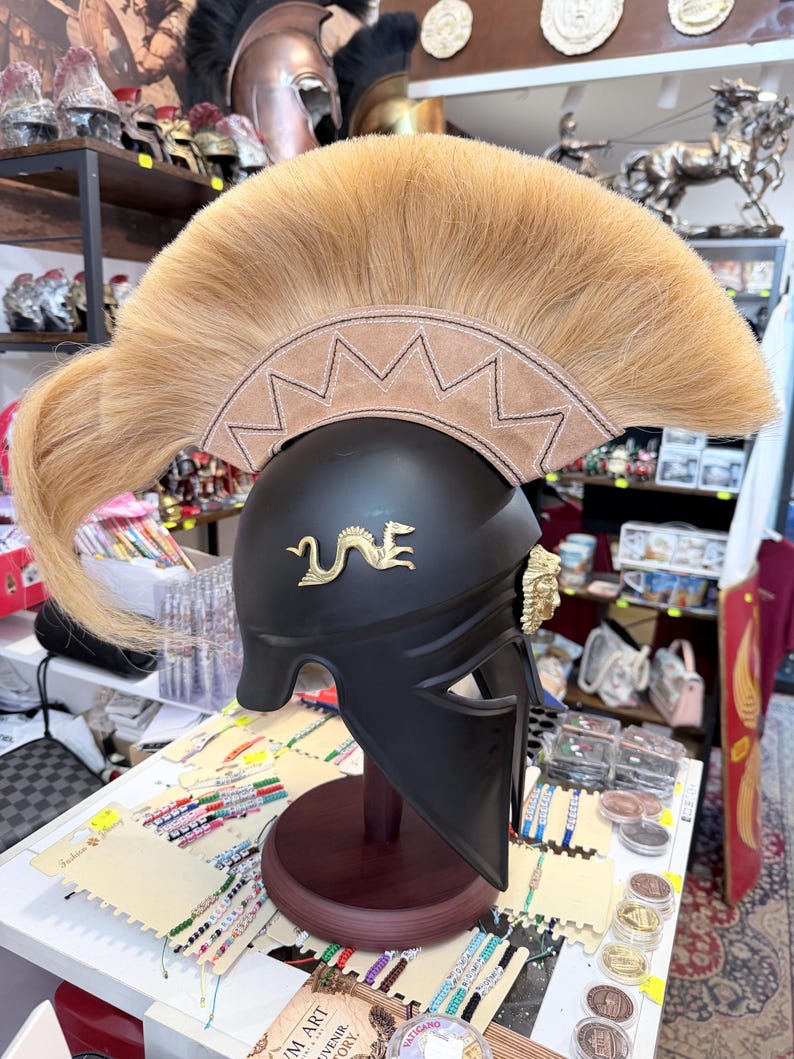 K&ouml;nnte beinhalten: Ein mattschwarzer Helm mit einem markanten, beigefarbenen Rosshaarkamm auf einer Basis mit Zickzack-N&auml;hten. Der Helm ist an der Seite mit einem goldenen Drachenemblem und in der N&auml;he des Gesichtsschutzes mit einem goldenen L&ouml;wenkopf verziert.
