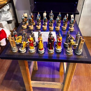 May include: A square chess board with a dark blue and black chequered pattern, featuring a set of hand-painted Roman-themed chess pieces. The pieces include soldiers, horses, and towers in various colours, with a certificate of authenticity in the centre.