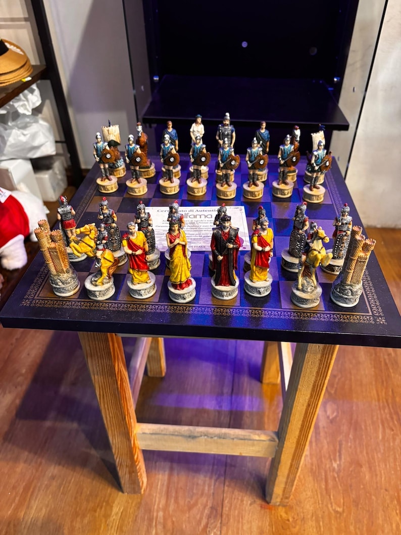 May include: A square chess board with a dark blue and black chequered pattern, featuring a set of hand-painted Roman-themed chess pieces. The pieces include soldiers, horses, and towers in various colours, with a certificate of authenticity in the centre.