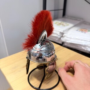 K&ouml;nnte beinhalten: Ein Miniaturhelm im r&ouml;mischen Stil mit einer polierten, silberfarbenen Metallkuppel, einem leuchtend roten B&uuml;rstenkamm, der von der Oberseite ausgeht, und zwei beweglichen Wangenklappen. Der Helm ist auf einem schwarzen Metallst&auml;nder mit rundem Fu&szlig; montiert.