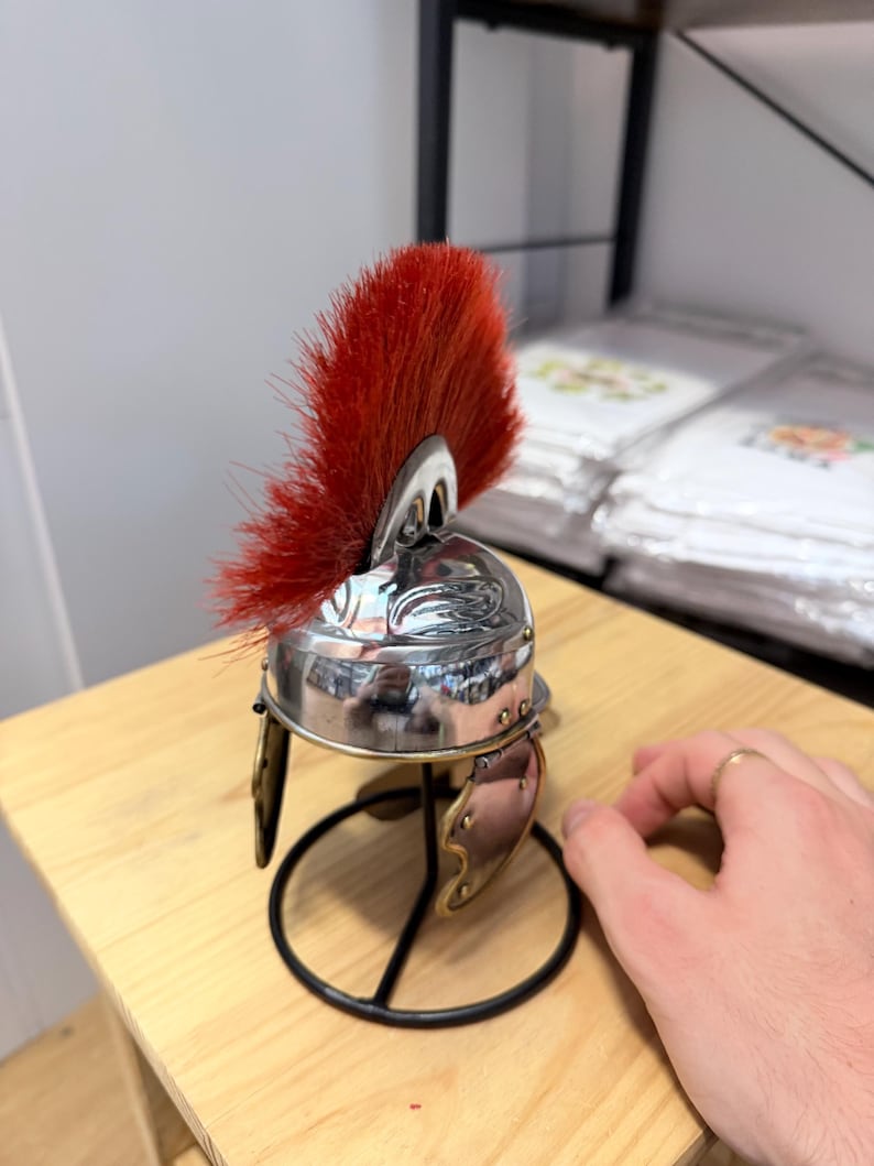 K&ouml;nnte beinhalten: Ein Miniaturhelm im r&ouml;mischen Stil mit einer polierten, silberfarbenen Metallkuppel, einem leuchtend roten B&uuml;rstenkamm, der von der Oberseite ausgeht, und zwei beweglichen Wangenklappen. Der Helm ist auf einem schwarzen Metallst&auml;nder mit rundem Fu&szlig; montiert.