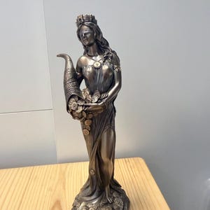 May include: A bronze-colored statue of a blindfolded figure wearing a crown and a draped gown. The figure holds a large cornucopia overflowing with gold-toned coins, with additional coins scattered around the base of the statue on the ground.