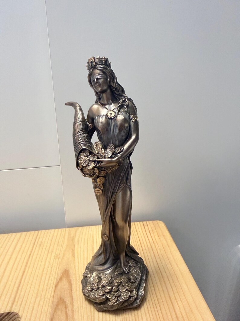 May include: A bronze-colored statue of a blindfolded figure wearing a crown and a draped gown. The figure holds a large cornucopia overflowing with gold-toned coins, with additional coins scattered around the base of the statue on the ground.
