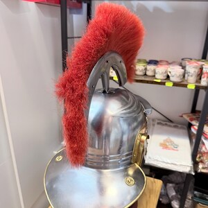Puede incluir: Un casco de metal plateado con una cresta alta y prominente de piel sint&eacute;tica de color rojo brillante que va de adelante hacia atr&aacute;s. El casco tiene un acabado liso y pulido con bordes dorados y peque&ntilde;os detalles circulares dorados.