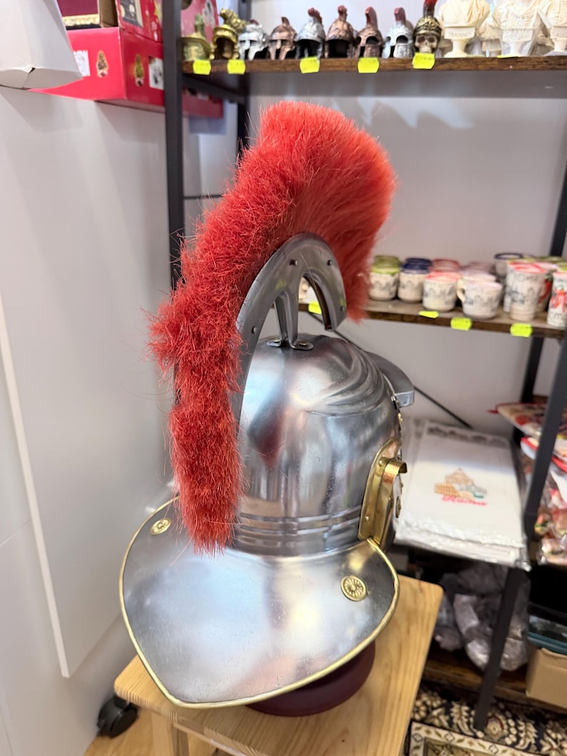 Puede incluir: Un casco de metal plateado con una cresta alta y prominente de piel sint&eacute;tica de color rojo brillante que va de adelante hacia atr&aacute;s. El casco tiene un acabado liso y pulido con bordes dorados y peque&ntilde;os detalles circulares dorados.