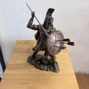 以下が含まれることがあります： 兜とマントを身につけたスパルタ戦士のブロンズ色の像。片手に槍、もう片方の手に丸い盾を持っている。盾には赤いラムダのシンボルが描かれ、3本の矢が突き刺さっている。台座にはLEONIDASと刻印されている。