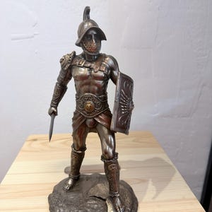 Pode incluir: Uma estatueta de gladiador de cor bronze, em p&eacute; sobre uma base texturizada. A figura est&aacute; vestida com uma armadura detalhada, incluindo um capacete, ombreiras e caneleiras. Ele segura uma pequena espada e um escudo. A base tem a inscri&ccedil;&atilde;o "GLADIATOR".
