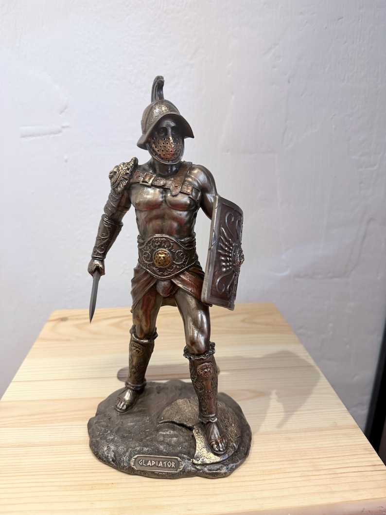 Pode incluir: Uma estatueta de gladiador de cor bronze, em p&eacute; sobre uma base texturizada. A figura est&aacute; vestida com uma armadura detalhada, incluindo um capacete, ombreiras e caneleiras. Ele segura uma pequena espada e um escudo. A base tem a inscri&ccedil;&atilde;o "GLADIATOR".