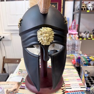 K&ouml;nnte beinhalten: Ein mattschwarzer Helm im r&ouml;mischen Stil mit einem goldenen Medusa-Emblem in der Mitte der Stirn, goldenen Seitenakzenten und einem beigefarbenen Kamm aus Kunsthaar, der auf einem dunklen Holzst&auml;nder zur Pr&auml;sentation montiert ist.
