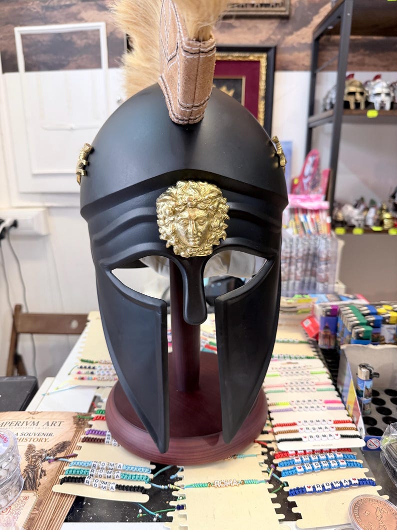 K&ouml;nnte beinhalten: Ein mattschwarzer Helm im r&ouml;mischen Stil mit einem goldenen Medusa-Emblem in der Mitte der Stirn, goldenen Seitenakzenten und einem beigefarbenen Kamm aus Kunsthaar, der auf einem dunklen Holzst&auml;nder zur Pr&auml;sentation montiert ist.