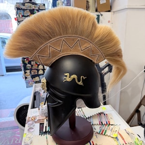 K&ouml;nnte beinhalten: Ein schwarzer Helm mit einem markanten beigefarbenen Kamm aus langen, steifen Borsten. Der Helm ist mit einem goldenen Drachenemblem an der Seite und einem goldenen L&ouml;wenkopfdetail in der N&auml;he des Wangenschutzes verziert und steht auf einem St&auml;nder.