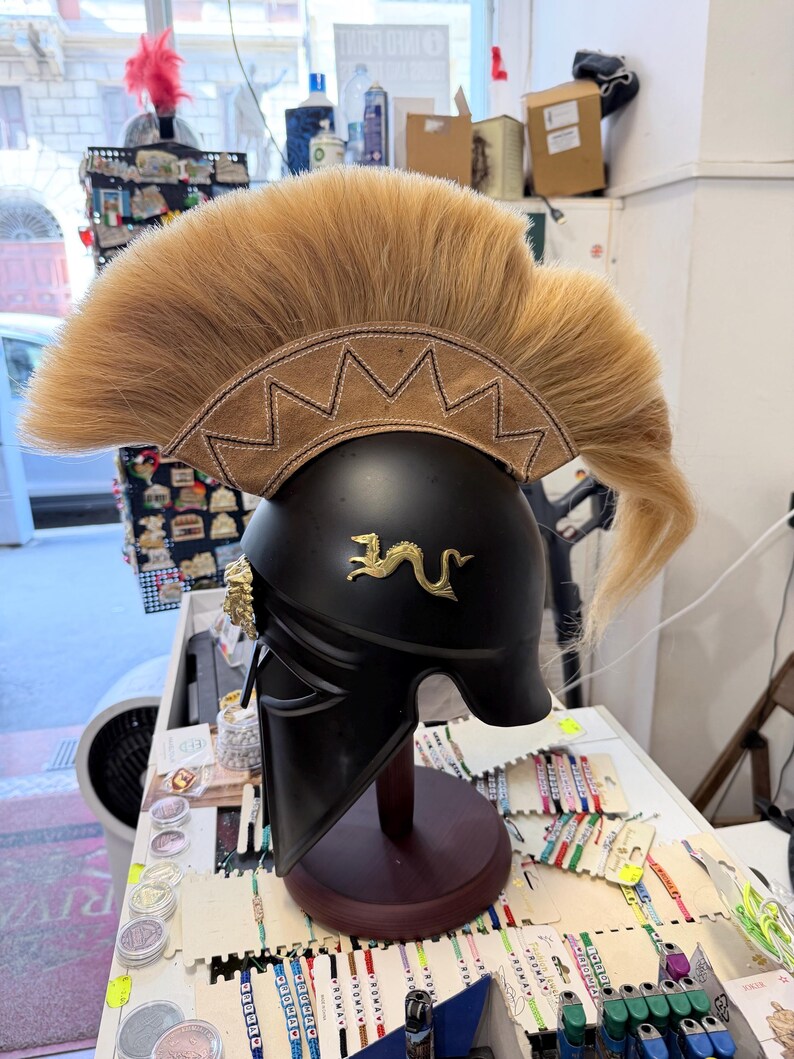 K&ouml;nnte beinhalten: Ein schwarzer Helm mit einem markanten beigefarbenen Kamm aus langen, steifen Borsten. Der Helm ist mit einem goldenen Drachenemblem an der Seite und einem goldenen L&ouml;wenkopfdetail in der N&auml;he des Wangenschutzes verziert und steht auf einem St&auml;nder.