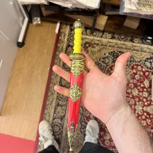 May include: A decorative Roman-style dagger with a yellow handle and a red sheath. The sheath is adorned with gold-colored embellishments and a circular emblem. The dagger is held in a person's hand.