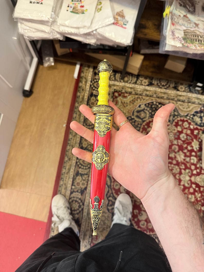 May include: A decorative Roman-style dagger with a yellow handle and a red sheath. The sheath is adorned with gold-colored embellishments and a circular emblem. The dagger is held in a person's hand.