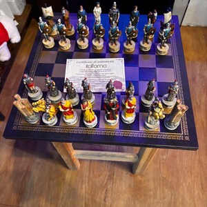 May include: A chess set featuring hand-painted Roman-themed pieces in blue and red colour schemes arranged on a dark square board. A white paper certificate of authenticity from Italfama is placed in the centre of the board among the chess pieces.
