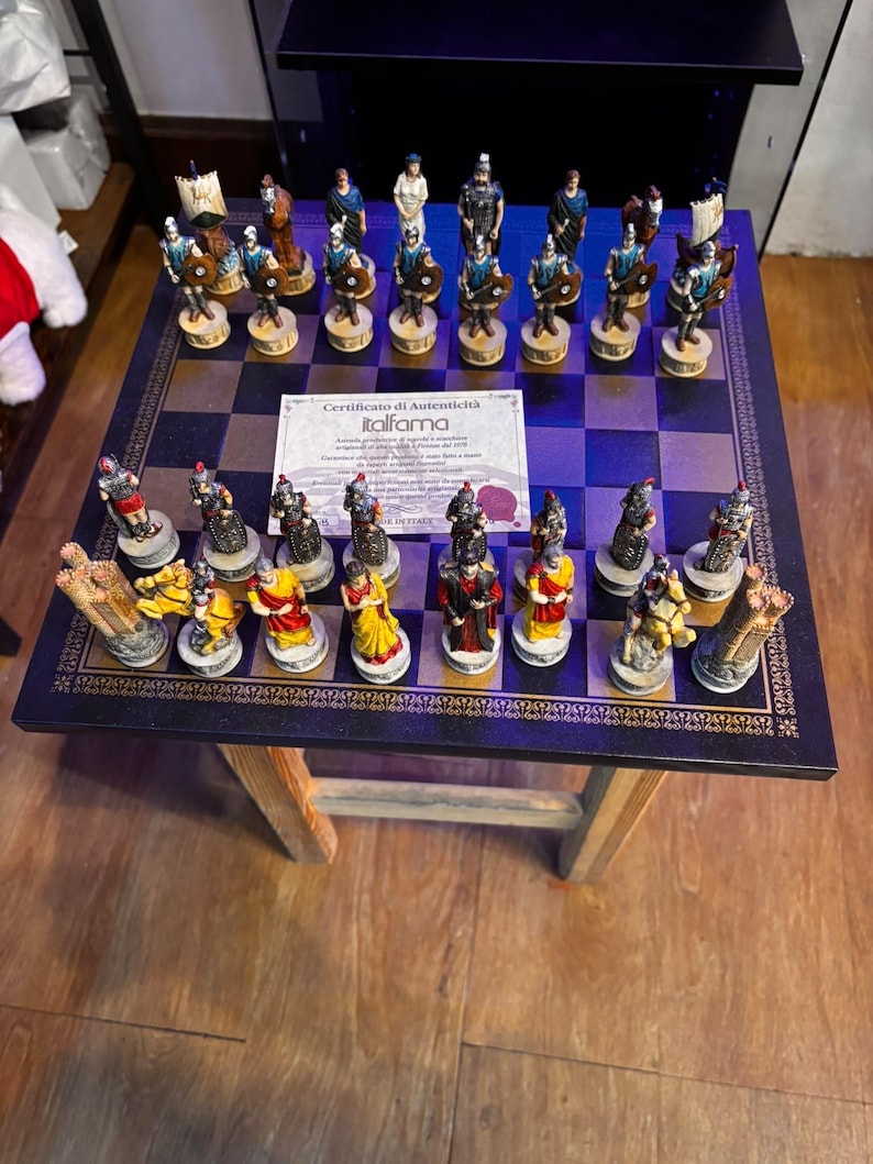 May include: A chess set featuring hand-painted Roman-themed pieces in blue and red colour schemes arranged on a dark square board. A white paper certificate of authenticity from Italfama is placed in the centre of the board among the chess pieces.