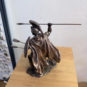 以下が含まれることがあります： 兜をかぶり、なびくマントをまとった戦士のブロンズ調のフィギュア。片手に長い槍、もう片方の手に丸い盾を持っています。盾には2本の矢が突き刺さっており、フィギュアは質感のある台座の上に立っています。