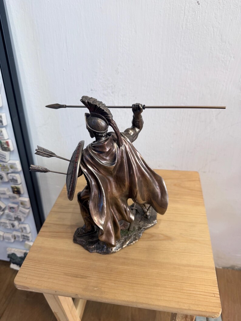 以下が含まれることがあります： 兜をかぶり、なびくマントをまとった戦士のブロンズ調のフィギュア。片手に長い槍、もう片方の手に丸い盾を持っています。盾には2本の矢が突き刺さっており、フィギュアは質感のある台座の上に立っています。