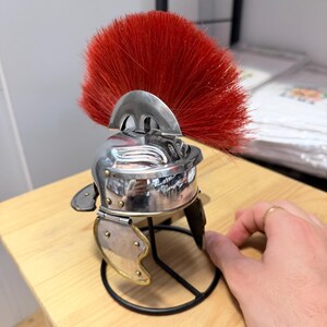 K&ouml;nnte beinhalten: Ein Miniaturhelm im r&ouml;mischen Stil aus silberfarbenem Metall mit einem markanten, leuchtend roten B&uuml;rstenkamm. Der Helm verf&uuml;gt &uuml;ber bewegliche Wangenklappen mit messingfarbenen Nieten und steht auf einem schwarzen Drahtst&auml;nder.