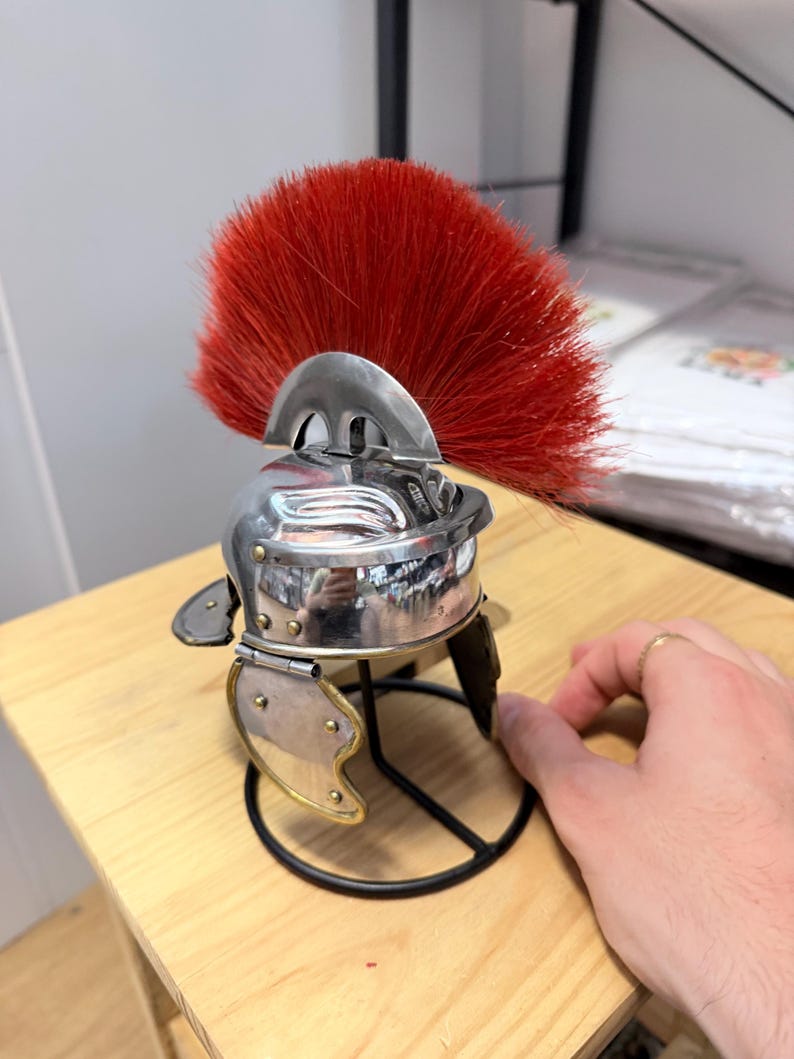 K&ouml;nnte beinhalten: Ein Miniaturhelm im r&ouml;mischen Stil aus silberfarbenem Metall mit einem markanten, leuchtend roten B&uuml;rstenkamm. Der Helm verf&uuml;gt &uuml;ber bewegliche Wangenklappen mit messingfarbenen Nieten und steht auf einem schwarzen Drahtst&auml;nder.