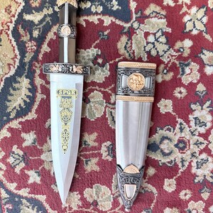 Peut inclure: Un poignard d&eacute;coratif avec une lame et un manche argent&eacute;s, des d&eacute;tails dor&eacute;s et une t&ecirc;te d'aigle sur le dessus. Le fourreau est argent&eacute; avec des bordures dor&eacute;es. Le poignard porte les lettres "SPQR" sur la lame.