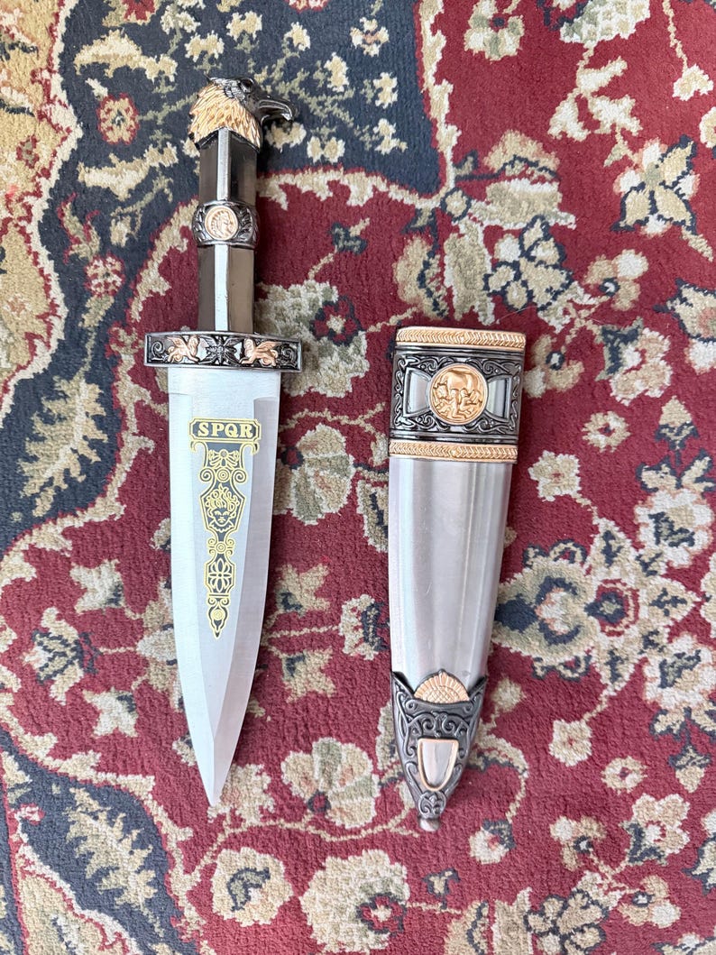 Peut inclure: Un poignard d&eacute;coratif avec une lame et un manche argent&eacute;s, des d&eacute;tails dor&eacute;s et une t&ecirc;te d'aigle sur le dessus. Le fourreau est argent&eacute; avec des bordures dor&eacute;es. Le poignard porte les lettres "SPQR" sur la lame.