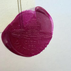 Pu&ograve; includere: Un sigillo in ceralacca color magenta con l'immagine in rilievo della Basilica di San Pietro e il testo CITTA DEL VATICANO. Il sigillo ha una forma arrotondata irregolare con una finitura liscia e lucida, impresso su carta chiara.