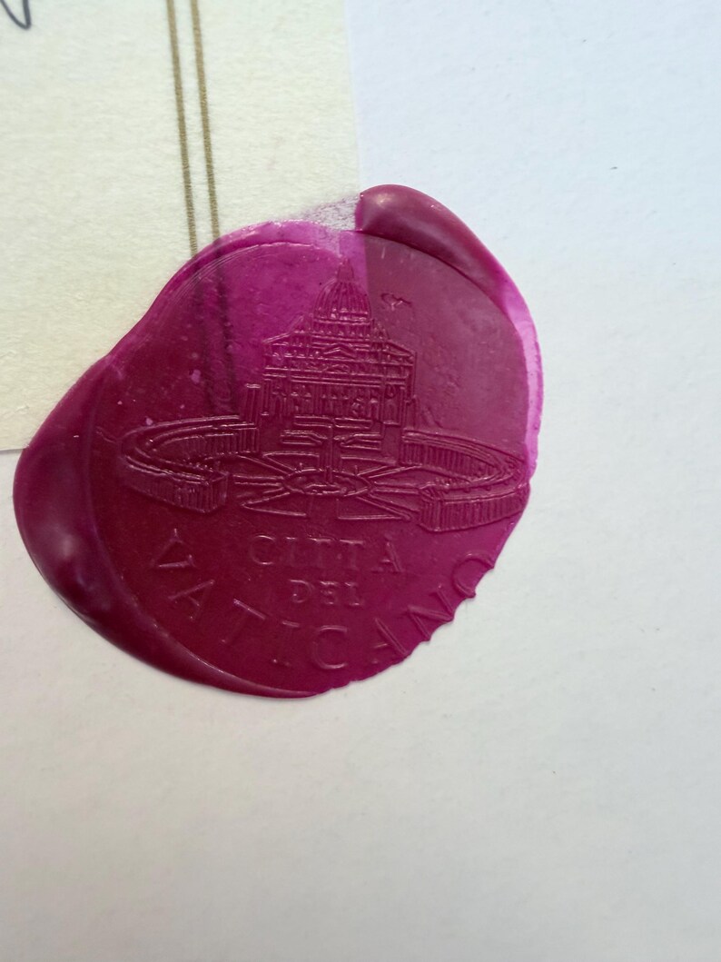 Pu&ograve; includere: Un sigillo in ceralacca color magenta con l'immagine in rilievo della Basilica di San Pietro e il testo CITTA DEL VATICANO. Il sigillo ha una forma arrotondata irregolare con una finitura liscia e lucida, impresso su carta chiara.