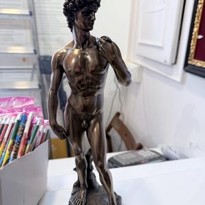 May include: Bronze-coloured statue of David, a classical male figure, standing on a textured base. The statue has detailed musculature and curly hair. The figure is posed with one hand resting on the shoulder.