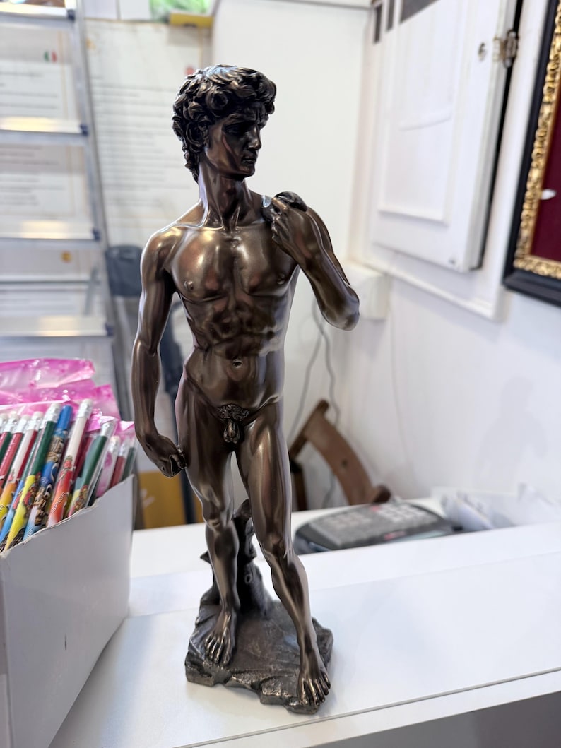 May include: Bronze-coloured statue of David, a classical male figure, standing on a textured base. The statue has detailed musculature and curly hair. The figure is posed with one hand resting on the shoulder.