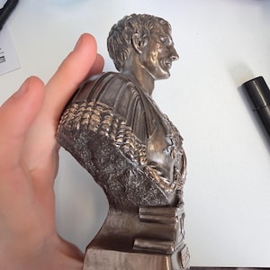 Peut inclure: Une sculpture buste de couleur bronze repr&eacute;sentant un personnage historique de profil, avec des traits du visage d&eacute;taill&eacute;s, des cheveux courts et un v&ecirc;tement textur&eacute; avec une cape drap&eacute;e. La figure est mont&eacute;e sur une base rectangulaire.