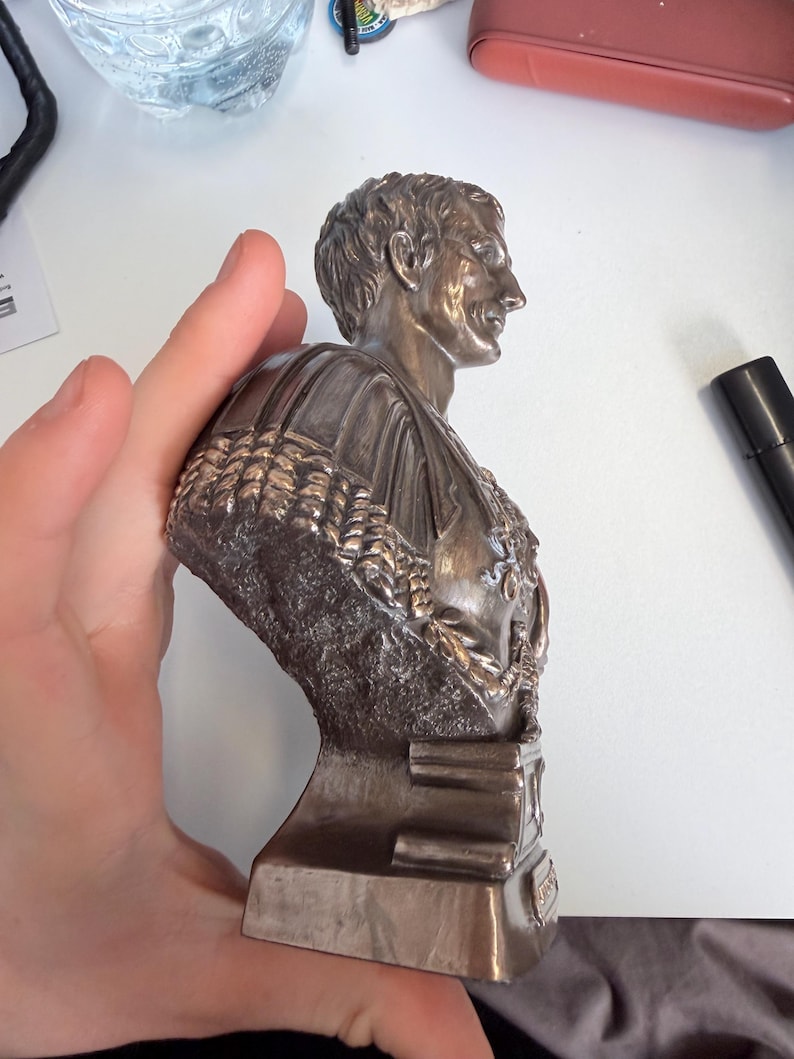 Peut inclure: Une sculpture buste de couleur bronze repr&eacute;sentant un personnage historique de profil, avec des traits du visage d&eacute;taill&eacute;s, des cheveux courts et un v&ecirc;tement textur&eacute; avec une cape drap&eacute;e. La figure est mont&eacute;e sur une base rectangulaire.