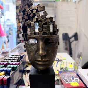 May include: A bronze-colored sculpture of a face with closed eyes and a fragmented top. The sculpture sits on a black rectangular base. The face has a serene expression, and the top of the head is made of small, square blocks.